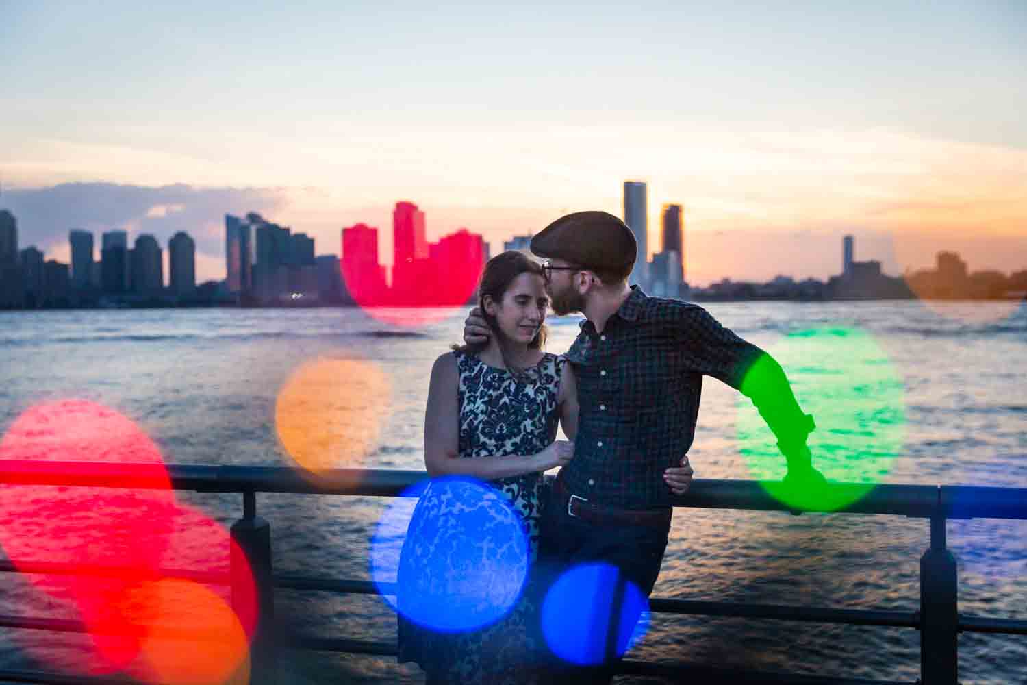 Man kissing woman's head with colored lights in front of Hudson River
