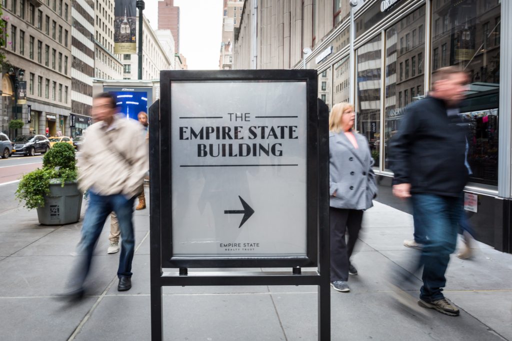 Empire State Building sign with people rushing past in a blur for an article on how to propose on top of the Empire State Building