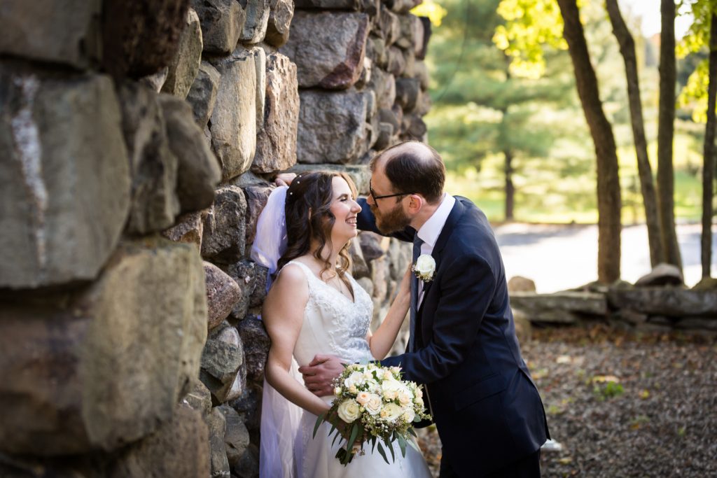 Bride leaning against stone wall and laughing with groom