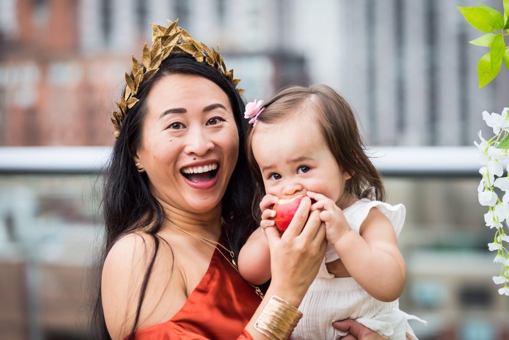 Birthday party photography of woman wearing gold laurel crown holding baby eating apple