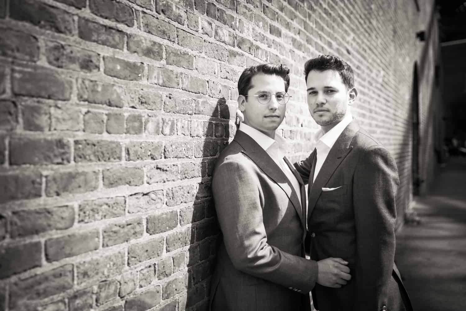 Black and white photo of two grooms leaning against brick wall