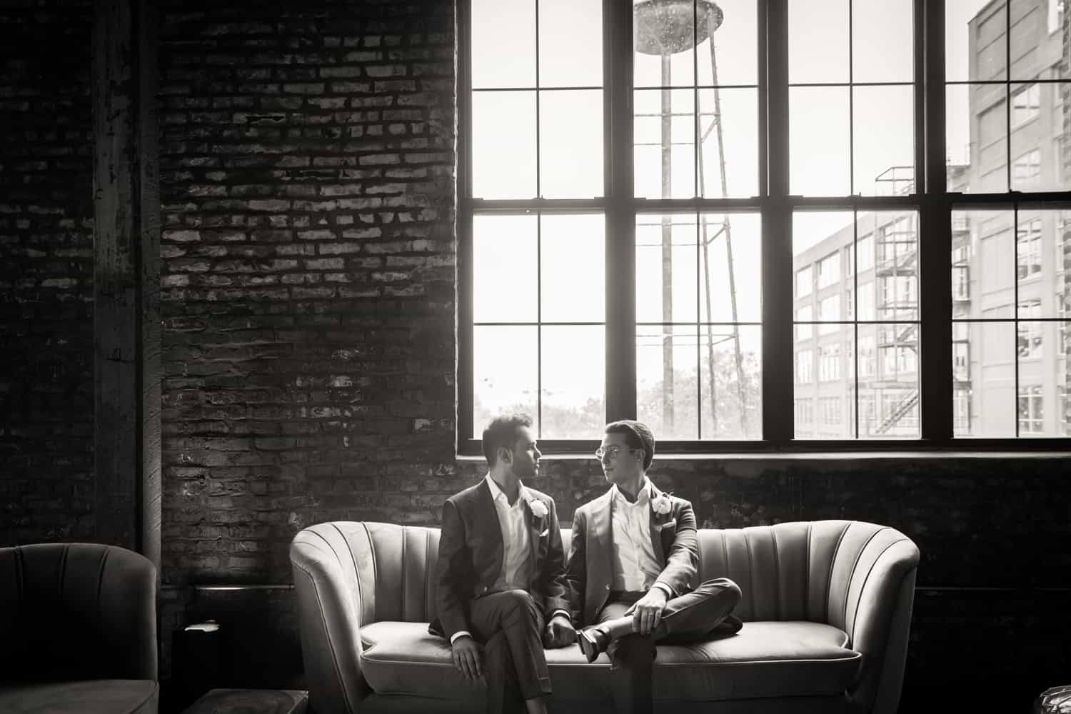 Black and white photo of two grooms sitting on couch under large window