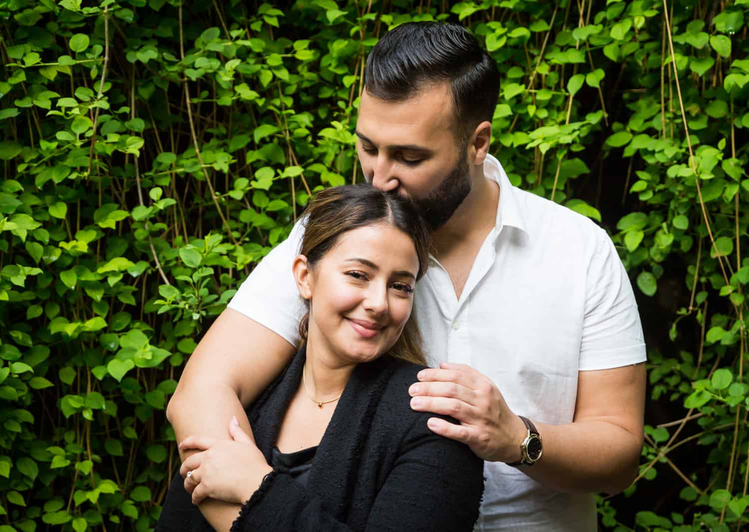 Man kissing woman's head in front of ivy-covered wall