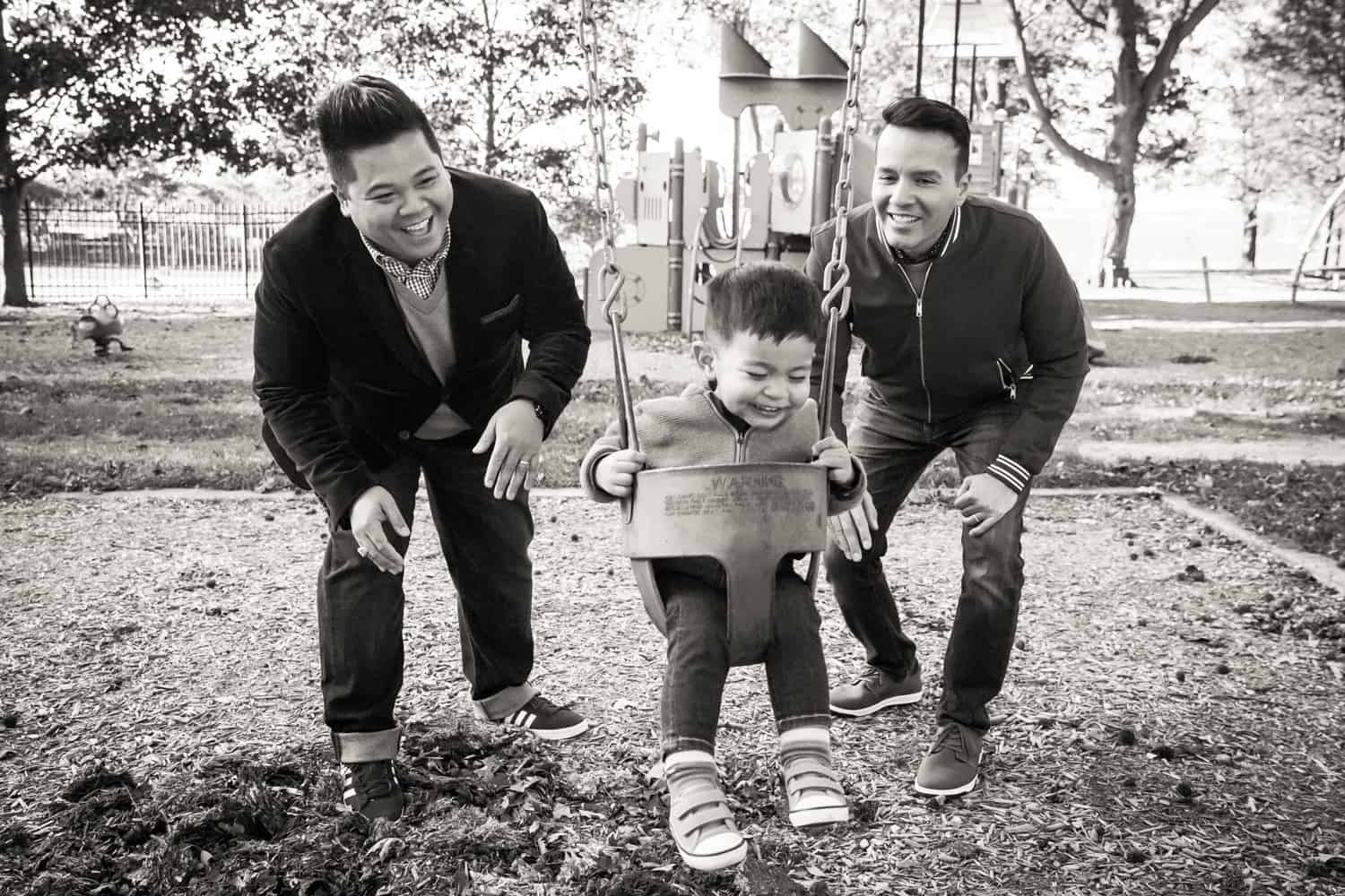 Black and white photo of two gay fathers with little boy on swing set