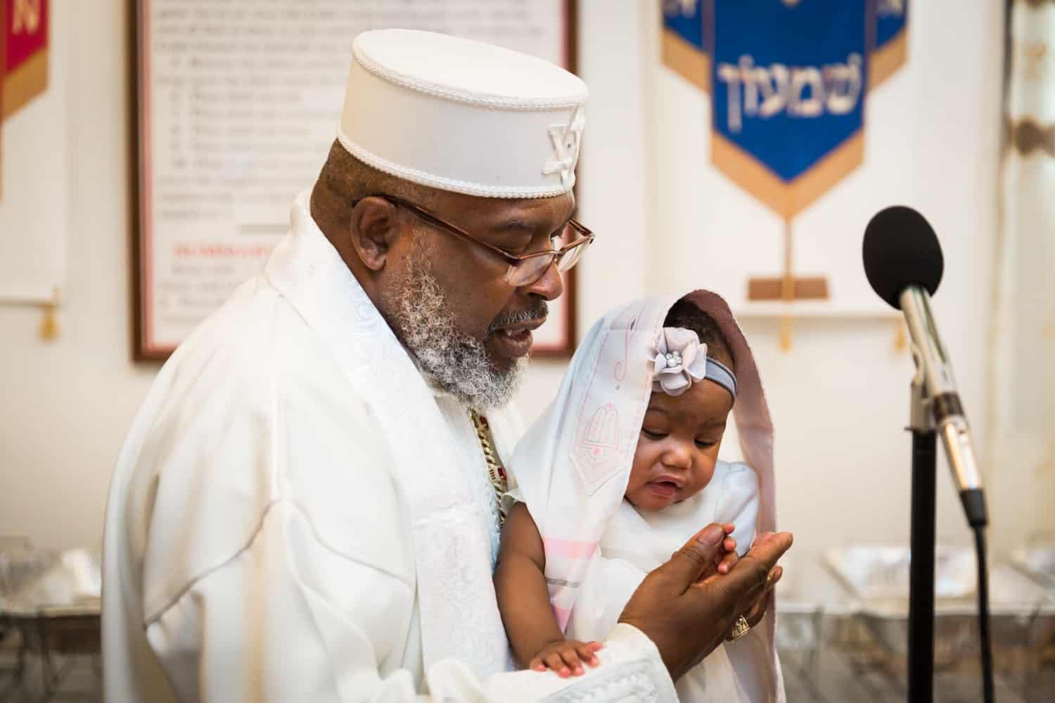 Pastor in white robe and hat holding little baby during Jamaica christening