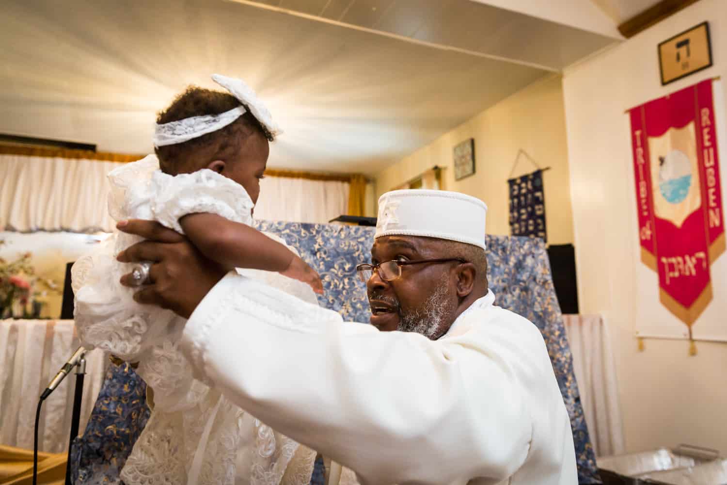 Pastor in white robes holding baby during Jamaica christening
