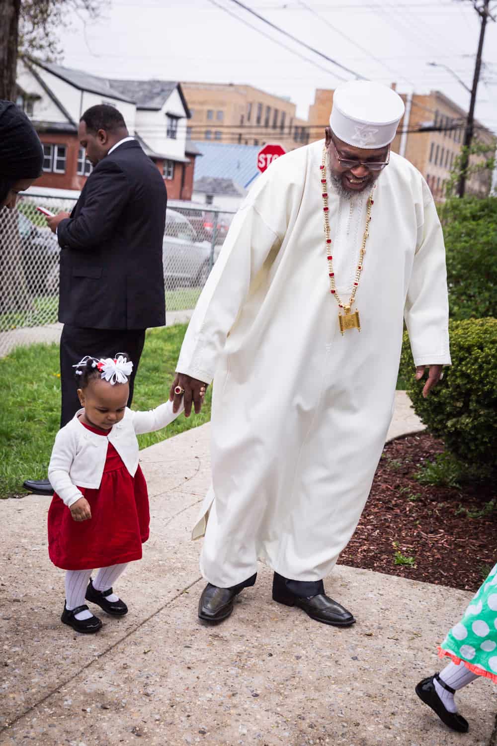 Pastor in white robe and hat holding little girl's hand on sidewalk