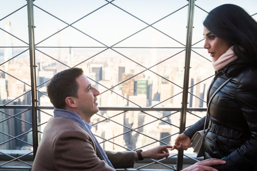 Man on one knee proposing to woman during proposal on top of the Empire State Building