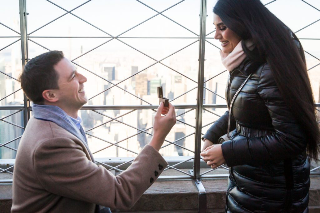 Man on one knee holding ring in front of woman during proposal on top of the Empire State Building