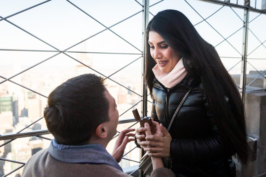 Woman looking down at man on one knee during proposal on top of the Empire State Building