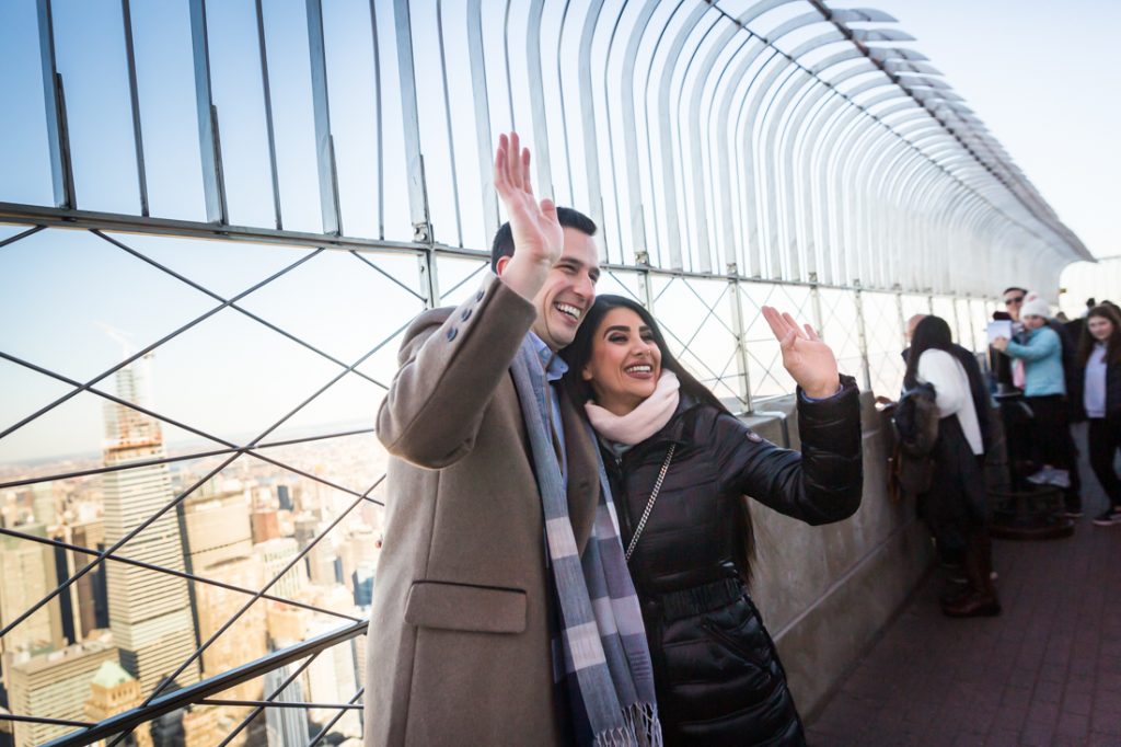 Couple waving to strangers in the window after proposal on top of the Empire State Building