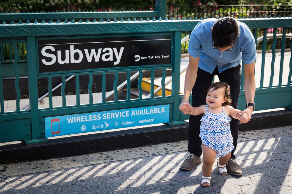 Father holding baby by hand in front of NYC subway sign