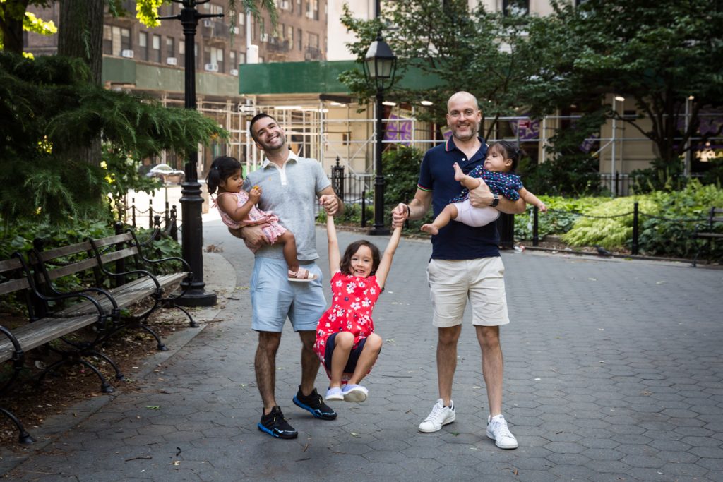 Two fathers swinging daughter by arms during a Washington Square Park family portrait