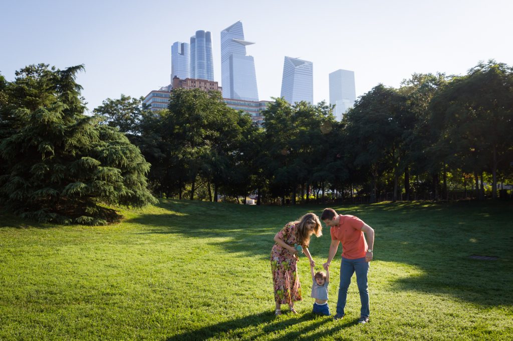 Parents holding little boy by the hand with NYC skyline during a Chelsea Waterside Park family portrait session