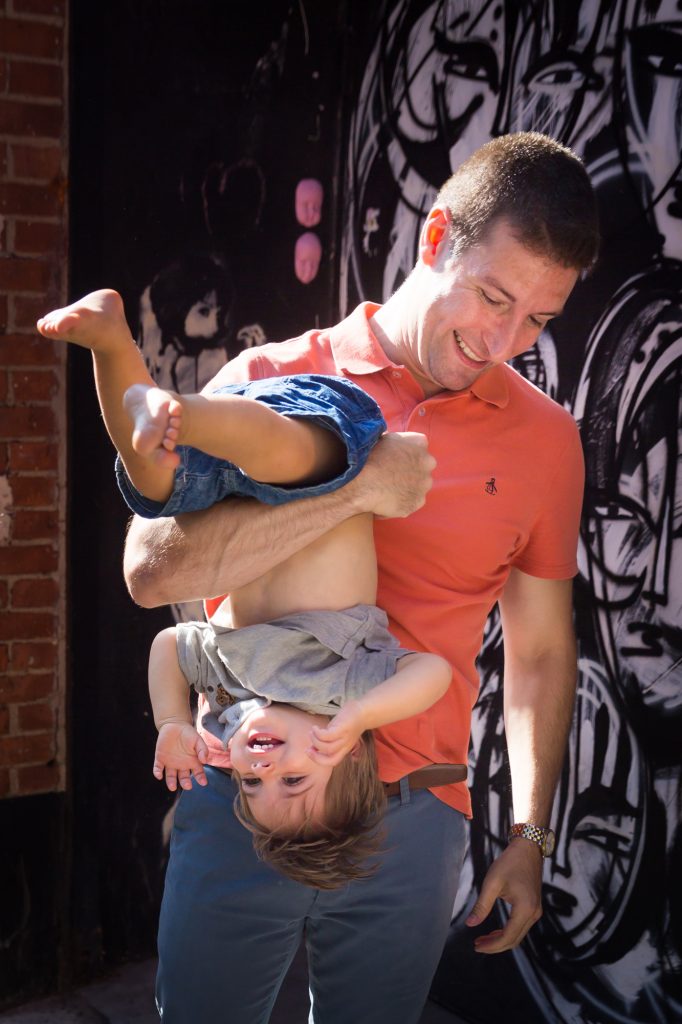 Father holding son upside down in front of black and white graffiti wall