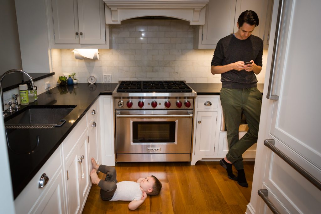 Young toddler with feet on cabinet and father looking at phone for an article on how to prepare for a newborn portrait session