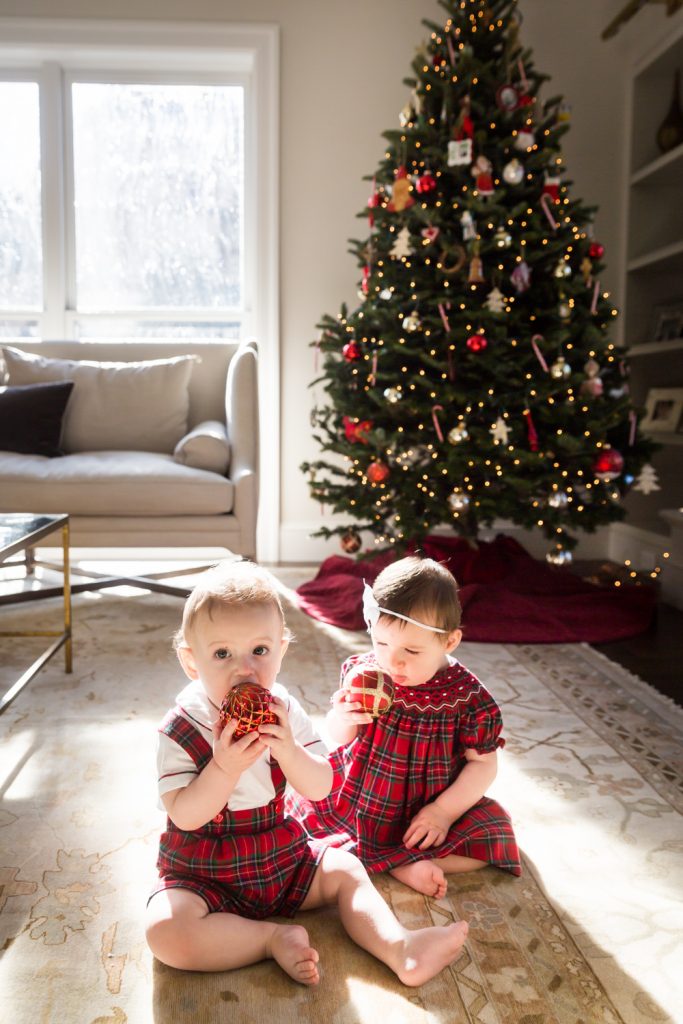 Two babies sitting on floor chewing on Christmas ornaments in front of tree