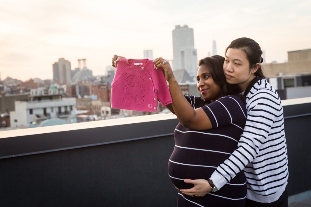 Couple hugging and holding up pink sweater during maternity portrait photo shoot