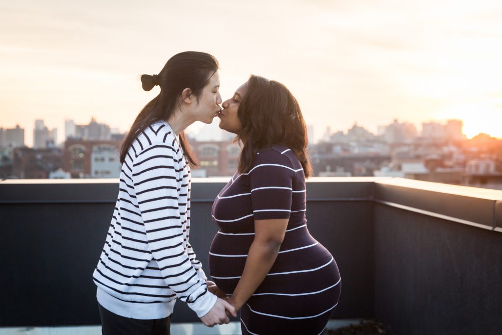 Couple holding hands and kissing on Brooklyn rooftop during maternity portrait photo shoot