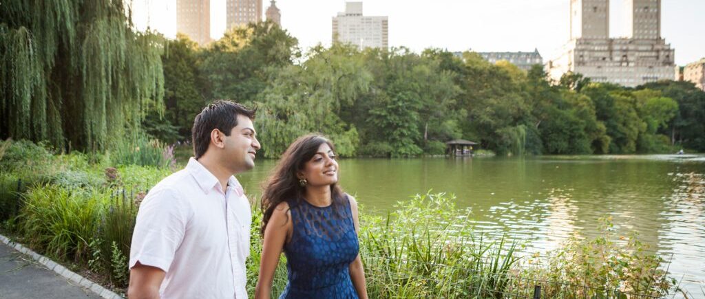 Couple walking past lake with NYC skyline in background during a Central Park engagement shoot