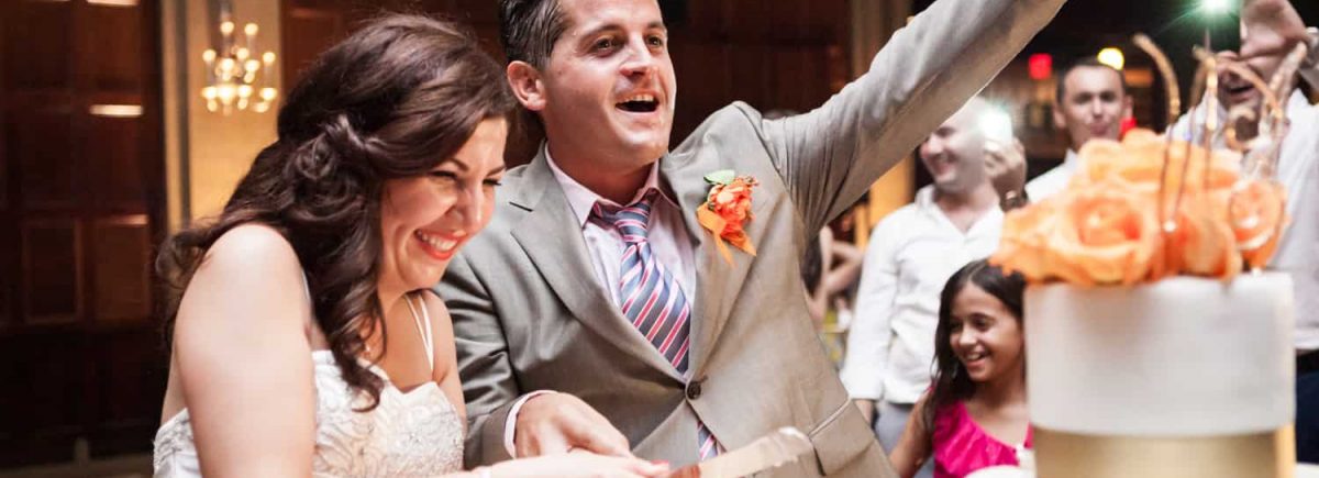 Groom cheers after cutting cake with bride at a Harvard Club wedding