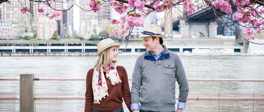Couple holding hands with cherry blossom branches overhead during a Roosevelt Island engagement portrait session