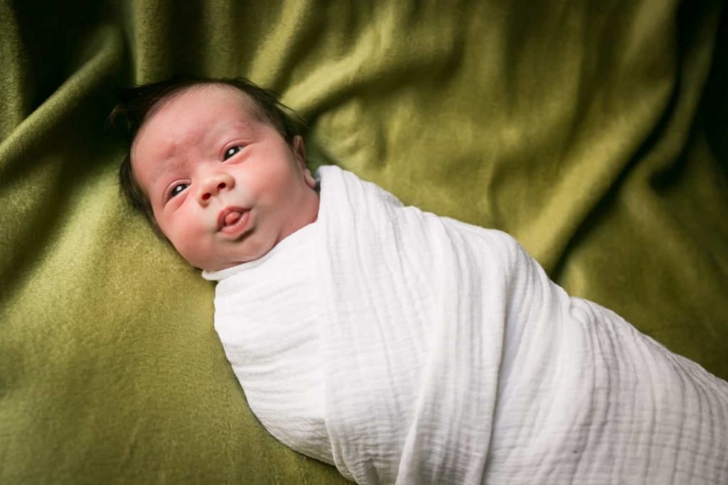 Swaddled newborn baby on green blanket for an article answering the question, 'is camera flash dangerous to newborns?'