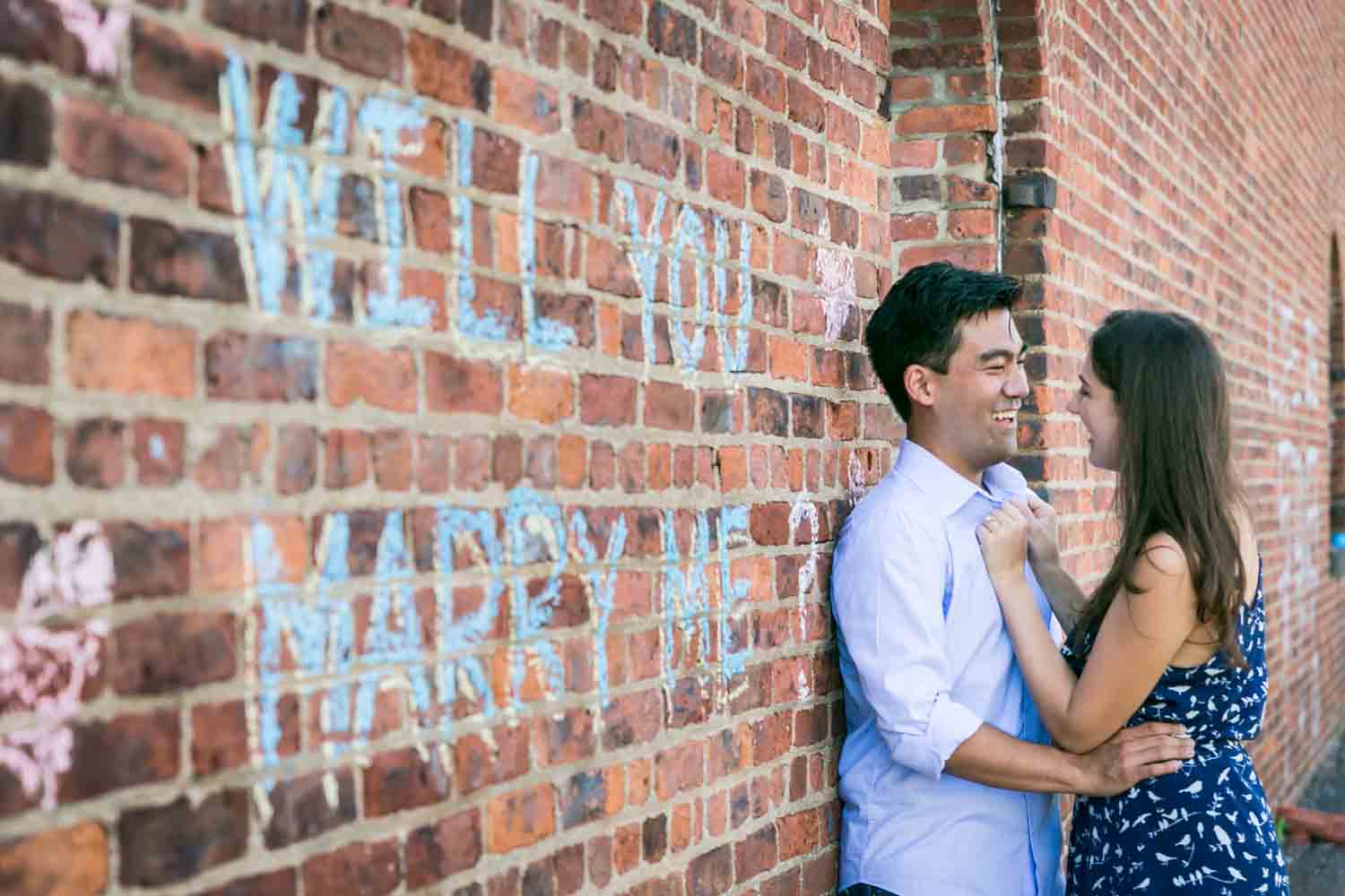 Couple leaning against brick wall with 'Will you marry me?' sign on it