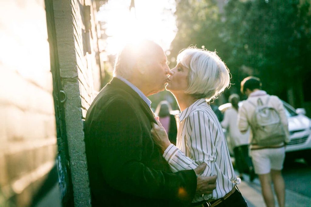 Photojournalistic portraits of older couple kissing against building