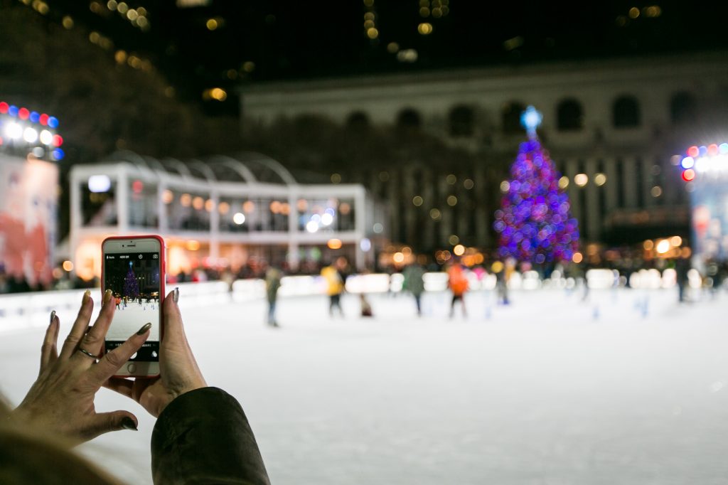Woman taking cell phone photo of ice skating rink for an article on NYC holiday card location suggestions