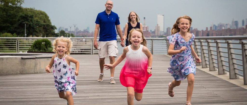Three little girls running in front of parents on boardwalk during a Gantry Plaza family portrait session