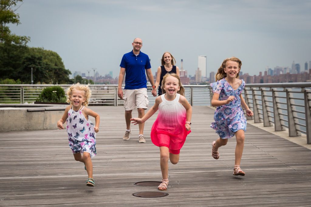 Three little girls running in front of parents on boardwalk during a Gantry Plaza family portrait session