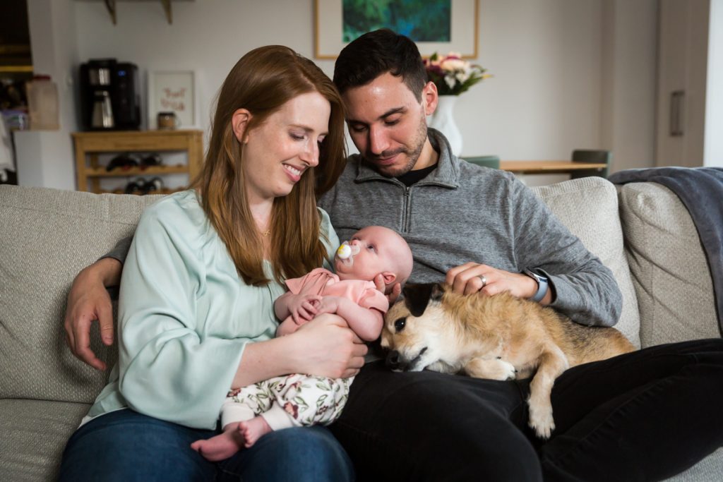 Parents sitting on couch and holding dog and newborn baby