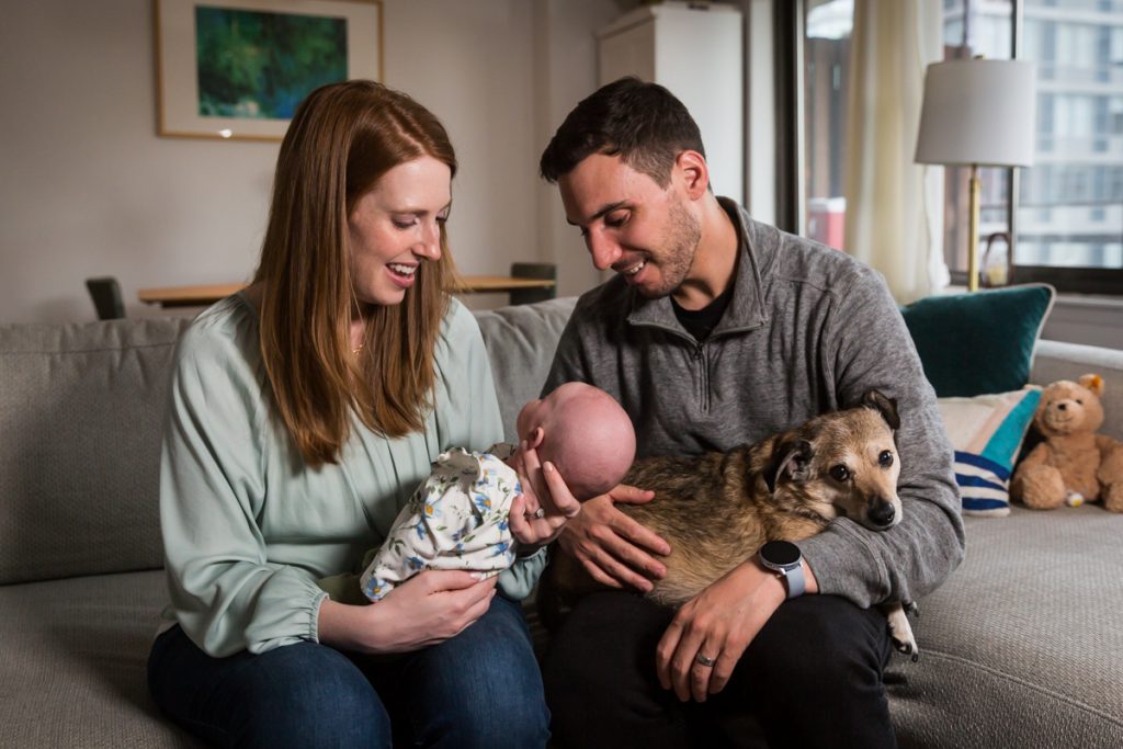Parents sitting on couch and holding dog and newborn baby