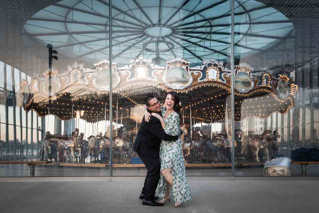 Couple laughing in front of Jane's Carousel during Brooklyn Bridge Park engagement shoot