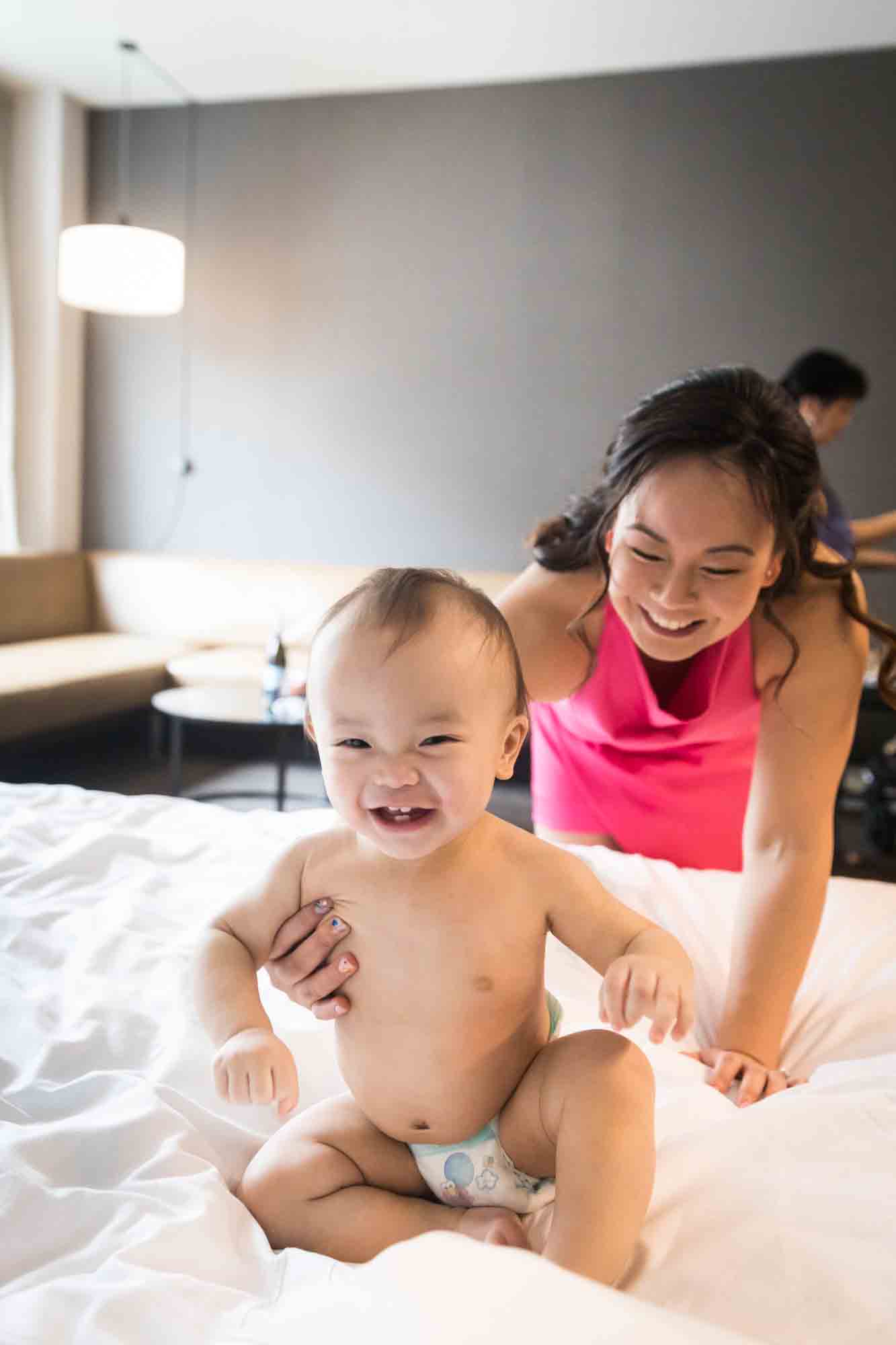 Mother grabbing naked baby sitting on bed