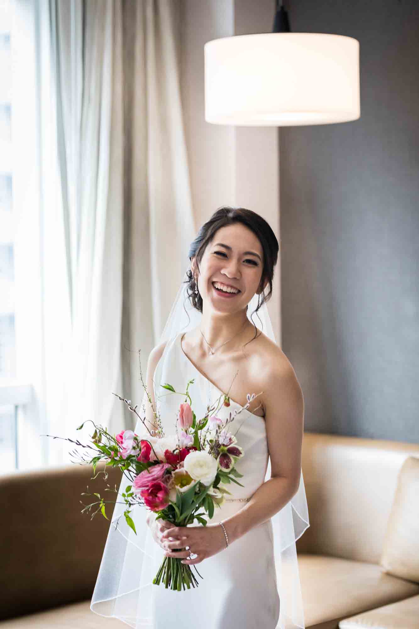 Bride laughing wearing one-shoulder dress and holding flowers