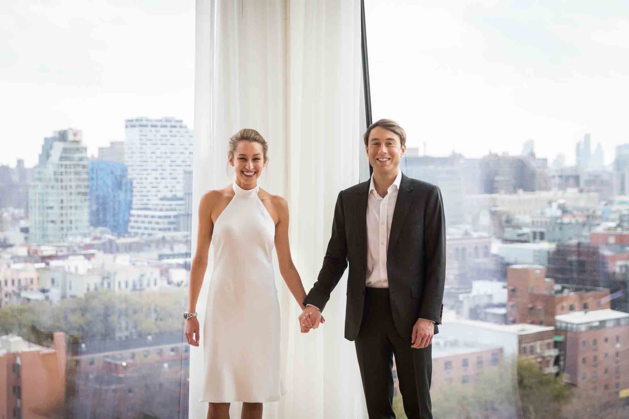 Bride and groom holding hands in front of windows for an article on how to plan a hotel room wedding ceremony