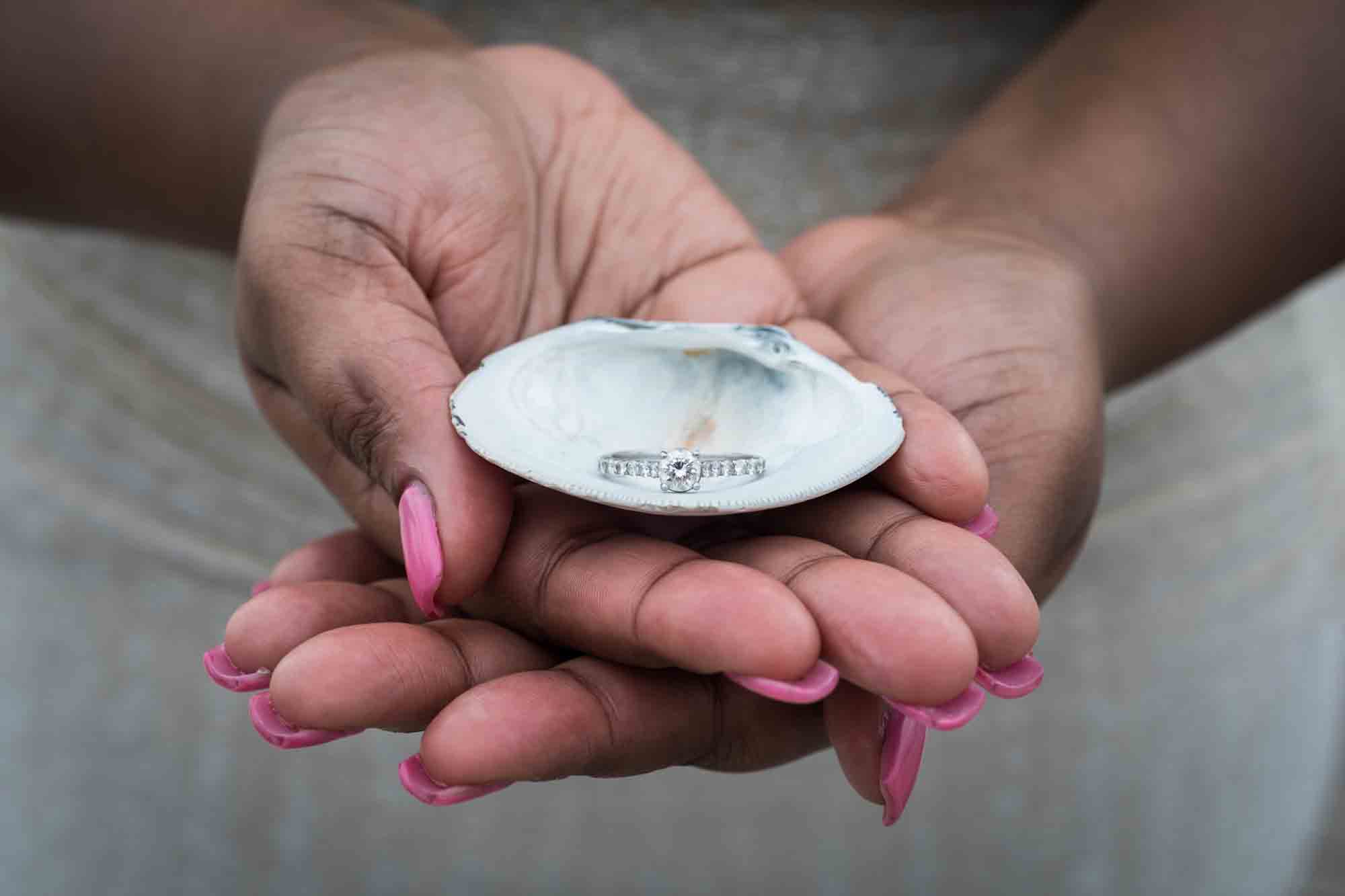Hands of a woman with pink fingernails holding a shell with an engagement ring