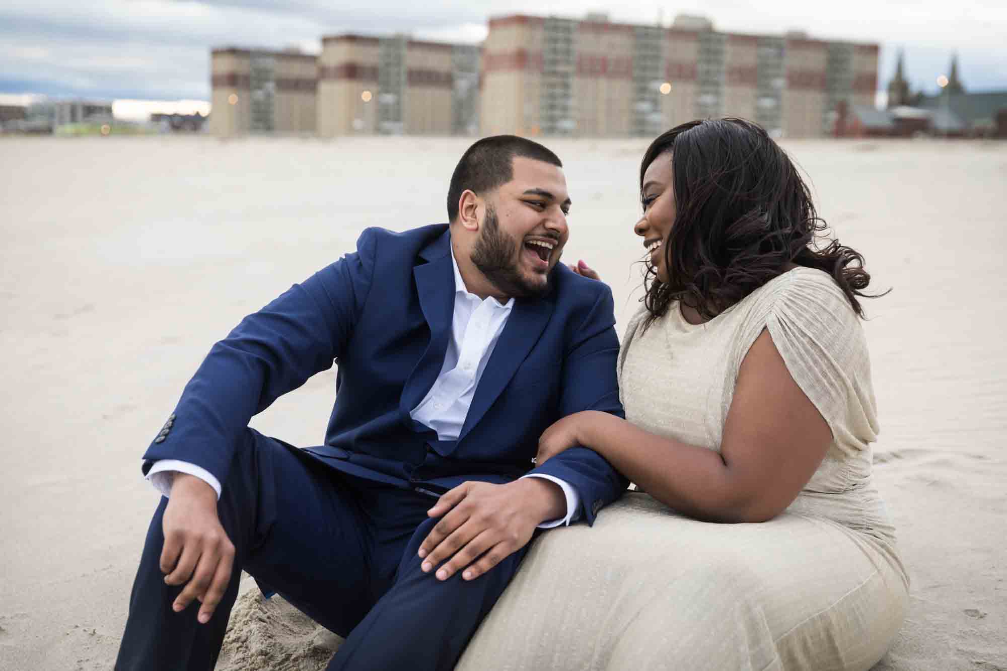 Couple sitting in the sand and laughing for an article on how to plan the perfect beach engagement photo shoot