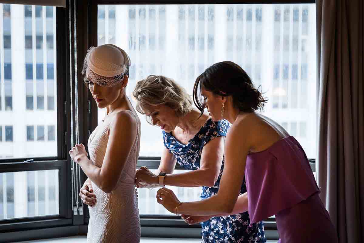 Mother and sister assisting bride get into dress in hotel room