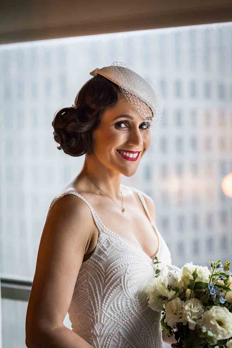 Smiling bride in front of window wearing vintage white hat with veil