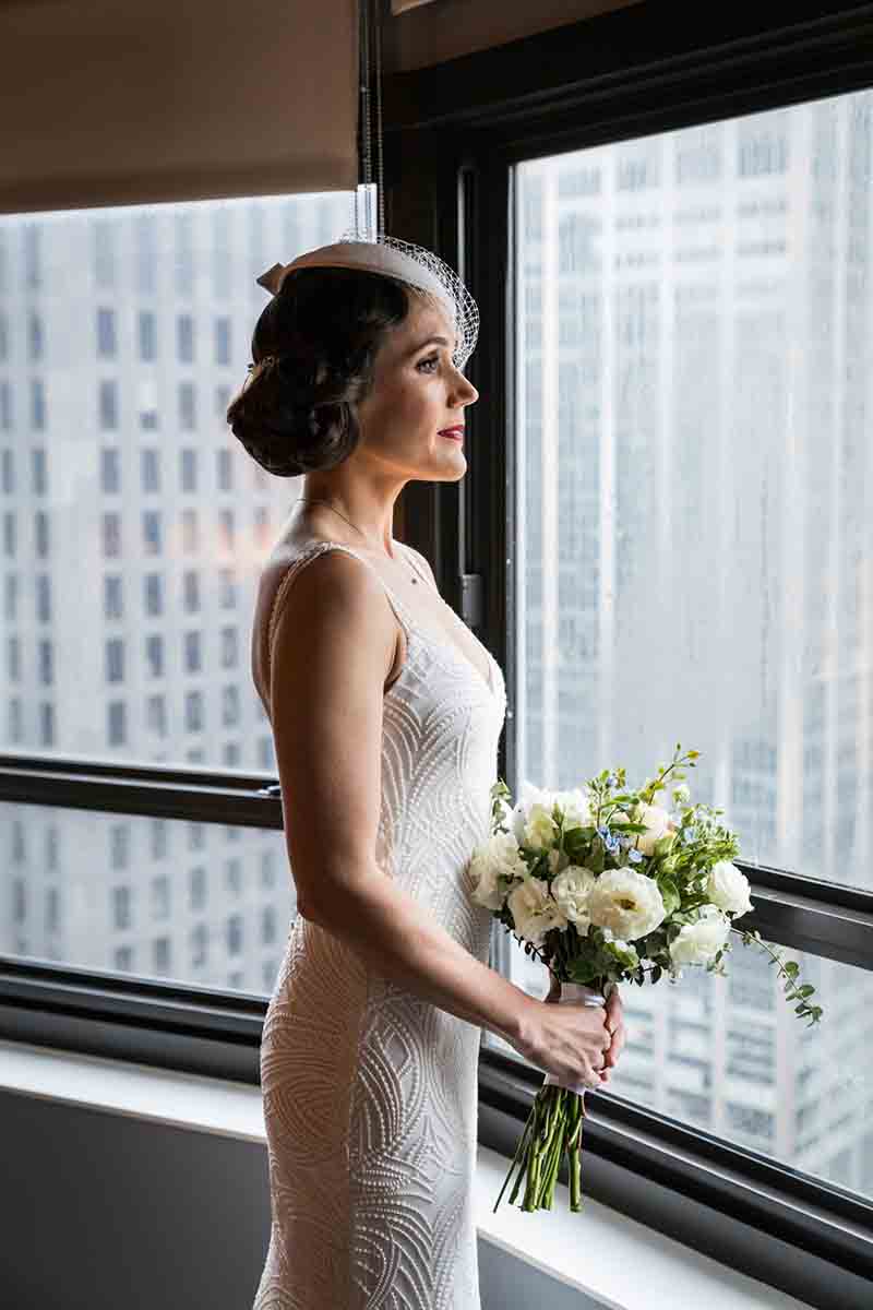 Bride looking out of window wearing vintage white hat with veil and holding bouquet