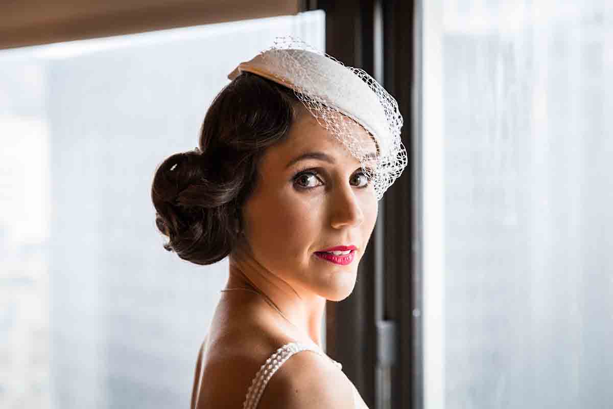 Bride in front of window wearing hair in a bun and white hat with veil