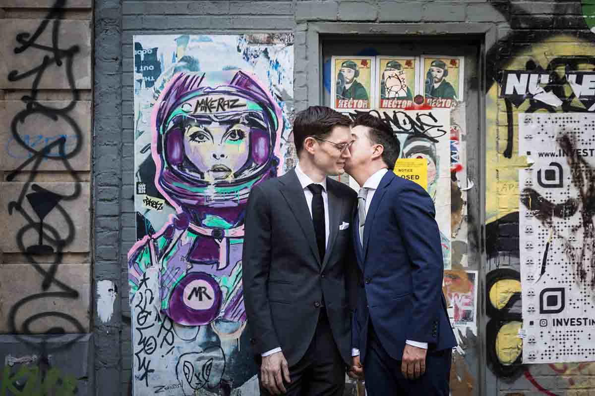 Man kissing another man on the cheek in front of graffiti wall