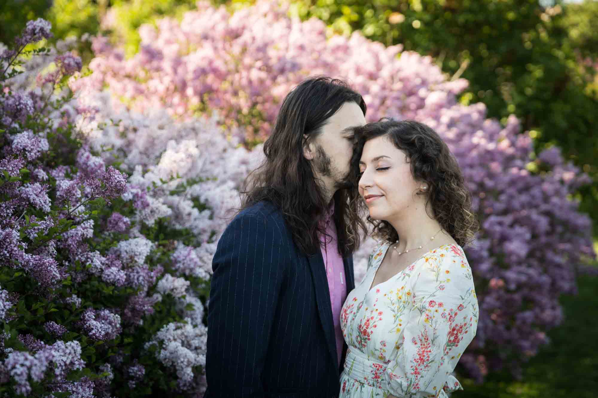Man kissing woman on side of head in front of lilac bushes