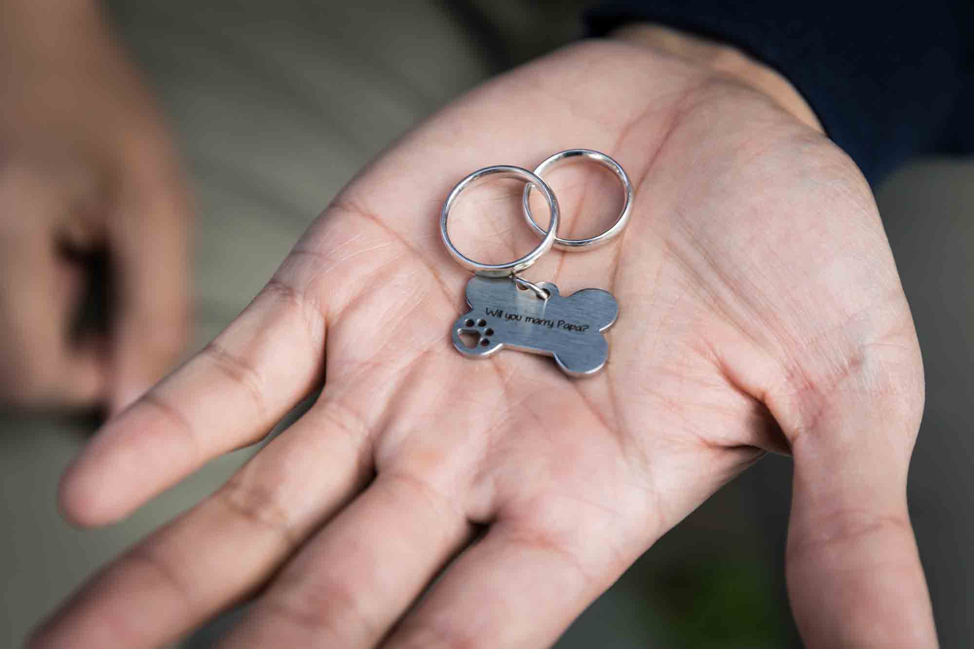 Man's palm holding two engagement rings and dog tag from a Central Park engagement photo shoot