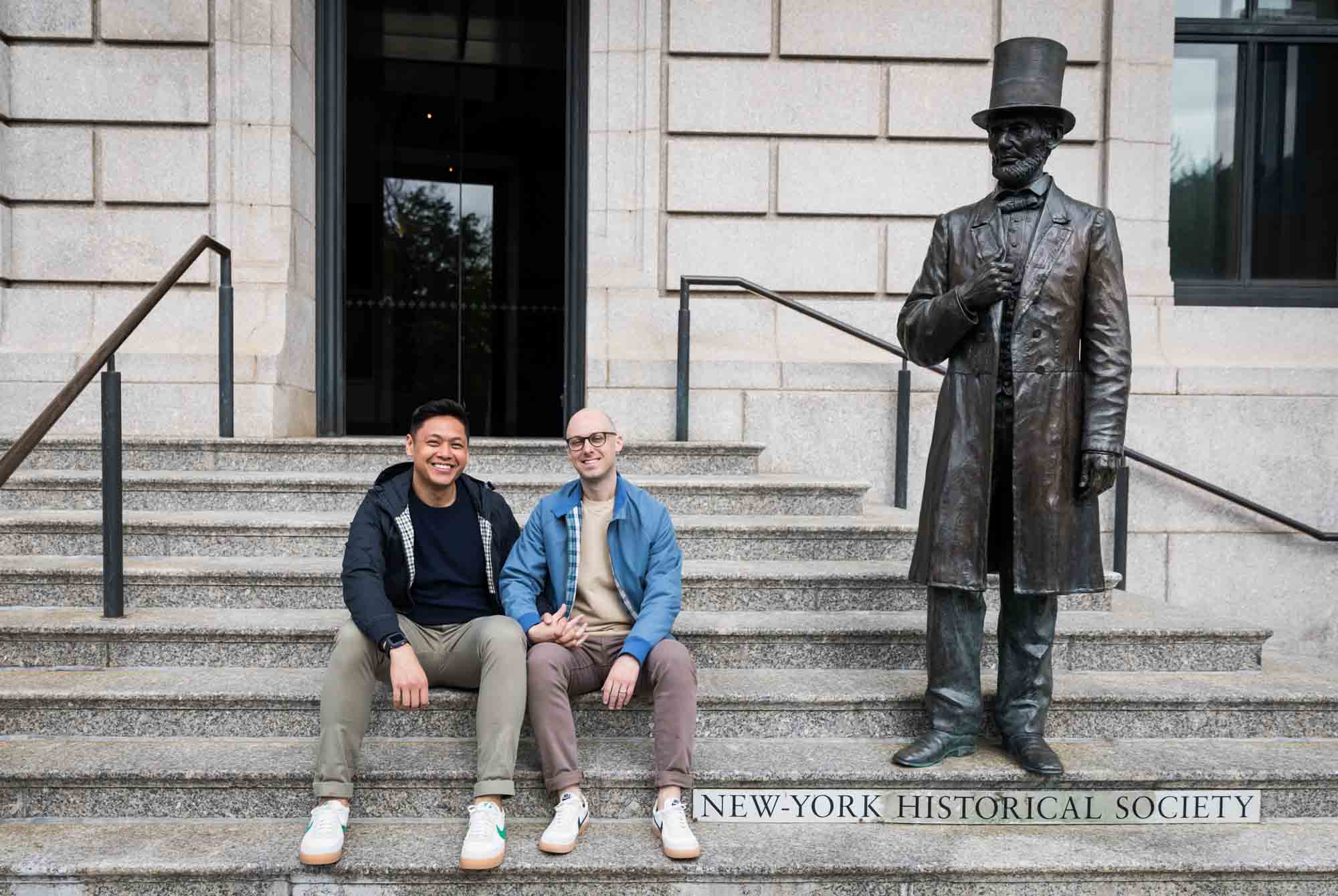Two men sitting on steps of New York Historical Society entrance with statue of Abraham Lincoln