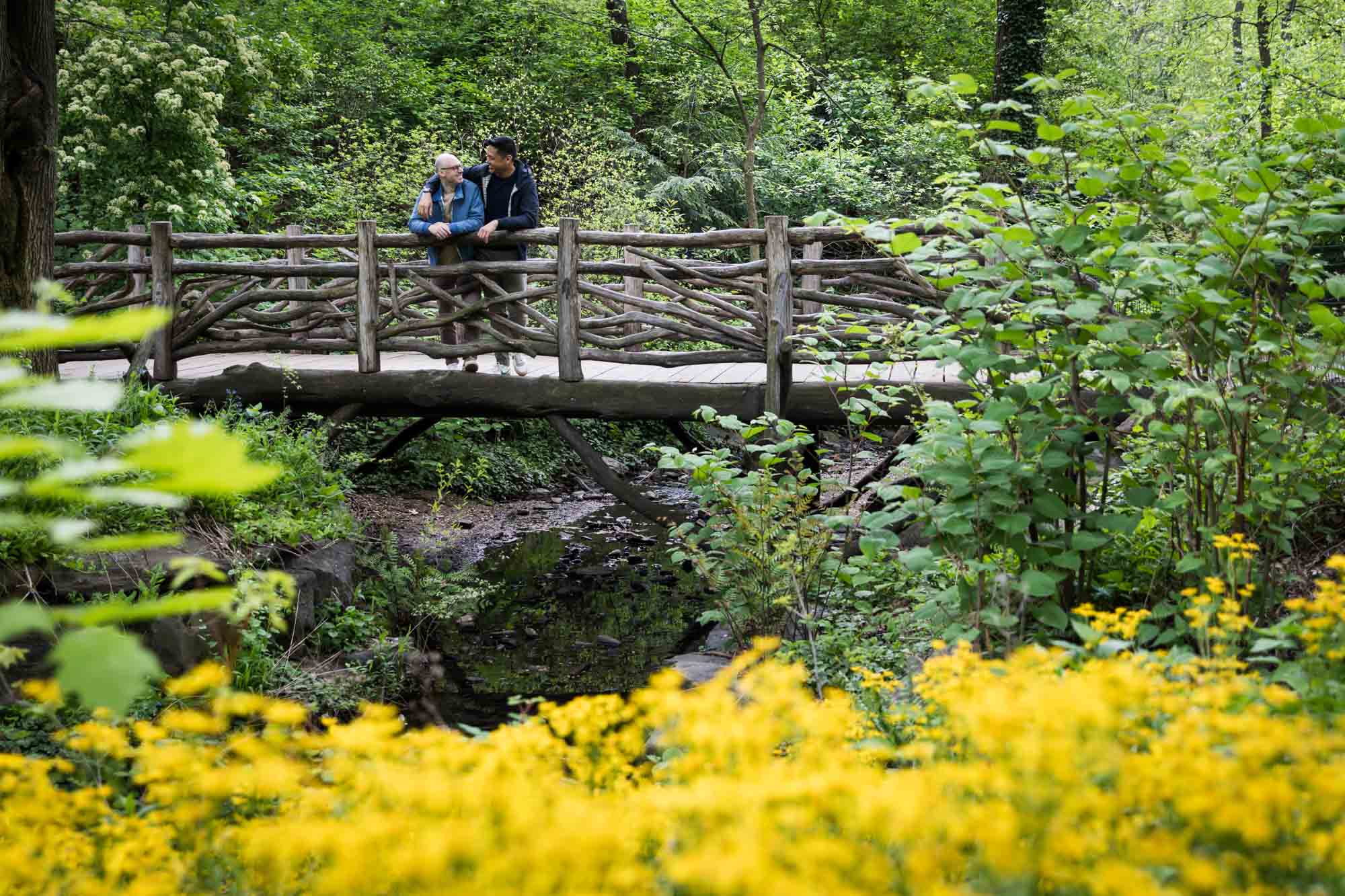 Two men standing on a wooden bridge with yellow flowers in the foreground from a Central Park engagement photo shoot