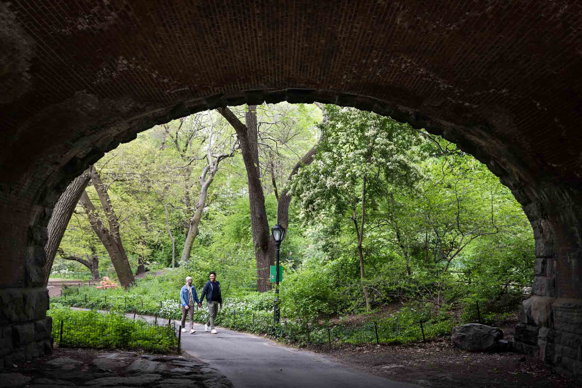 Two men walking on pathway under bridge from a Central Park engagement photo shoot